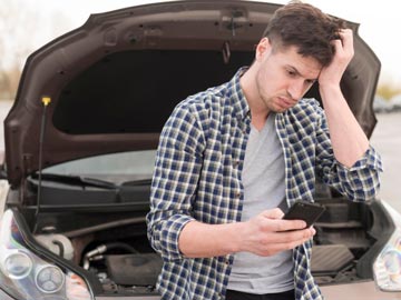 Eine Person in einem karierten Hemd sieht gestresst aus, während sie ihr Handy neben einer offenen Motorhaube überprüft, was auf Autoprobleme hinweist.